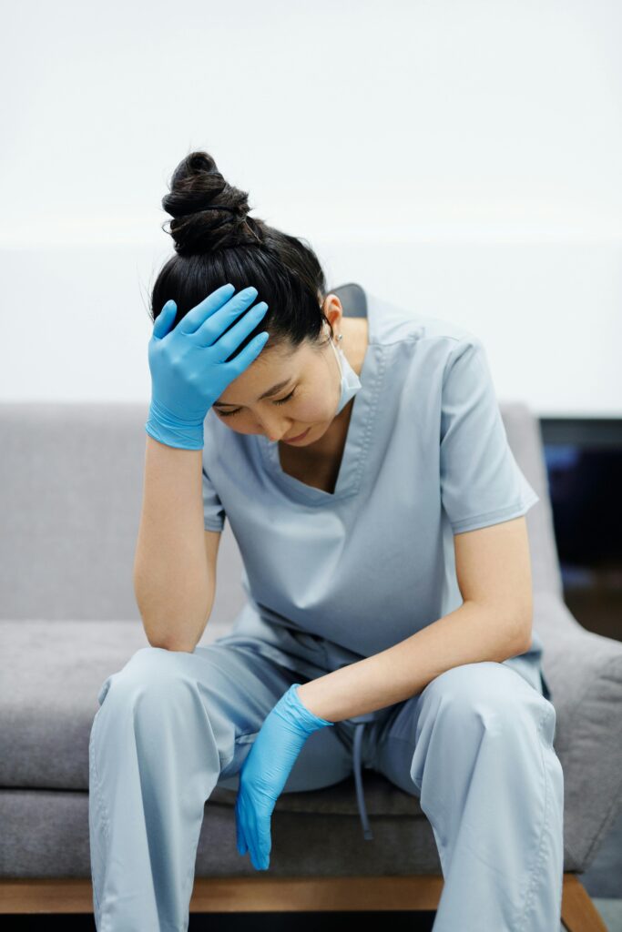 A tired female healthcare worker in scrubs sits on a couch with her head in her hand, looking stressed.